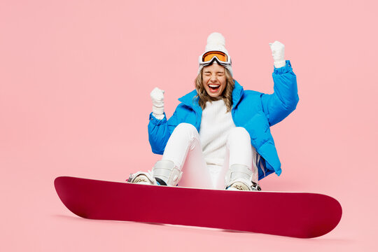 Bottom view snowboarder woman wear blue suit goggles mask hat ski padded jacket sit do winner gesture isolated on plain pastel pink background. Winter extreme sport hobby weekend trip relax concept.