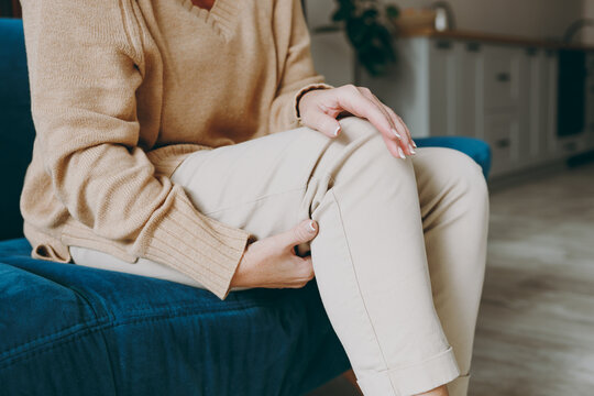 Cropped close up photo of pensioner woman wearing casual clothes sit on blue sofa hold knee suffer from leg knee pain inflammation stay home flat spend free spare time in living room indoor grey wall.