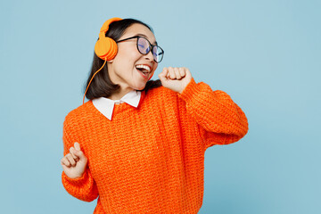 Young woman of Asian ethnicity in orange sweater glasses headphones listen to music raise up hands dance have fun on party sing song in microphone isolated on plain pastel light blue cyan background