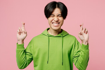 Young man of Asian ethnicity wears green hoody waiting for special moment, keeping fingers crossed,...