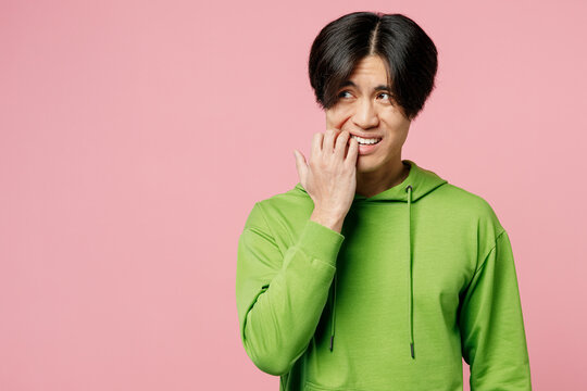 Young mistaken sad disappointed man of Asian ethnicity in green hoody look aside biting nails fingers say oops isolated on plain pastel light pink background studio portrait People lifestyle concept