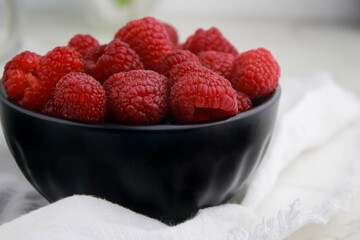 close up of fresh raspberries in dark bowl, blurred white background 