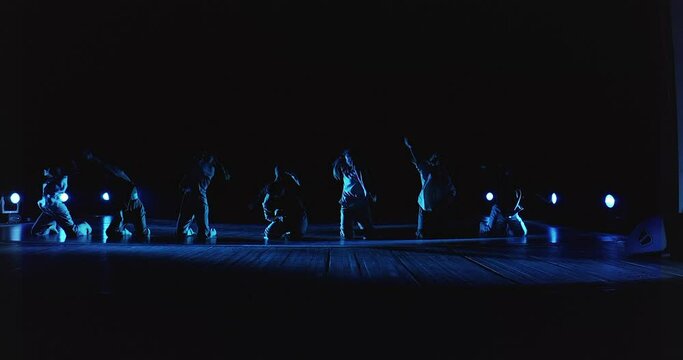 A group of dancers perform on stage. A group of artists are rehearsing on stage with lights and smoke. Girls dance in a performance on stage.