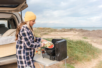 woman traveler prepares food on portable gas stove, on a folding table in a tranquil coastal spot...
