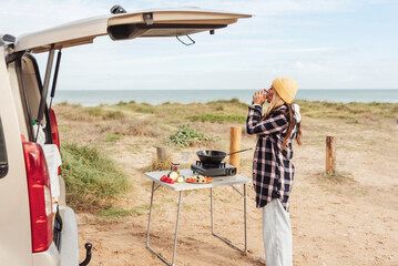 woman traveler prepares food on portable gas stove, on a folding table in a tranquil coastal spot outside of her camper van