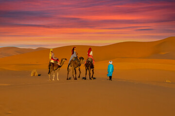 Three Riders And Their Handler Travel Through The Saharan Desert On Their Camels In Morocco - obrazy, fototapety, plakaty