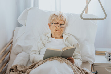 Sick senior woman lies in a hospital bed and reads a book - 557262702
