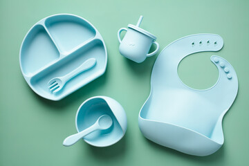 Silicone tableware set on color background. Flat lay baby bib, bowl with spoon, fork, cup. Baby feeding concept.