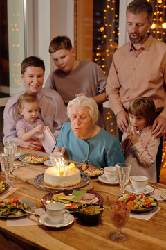 Portrait Of Grandma Blowing Out Cake Candles During Her Birthday Party In Bosom Of Her Family Vertical Shot