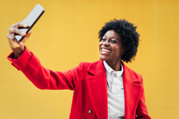 Afro woman smiling while taking selfies with her mobile phone against a yellow wall. Technology and social media concept.