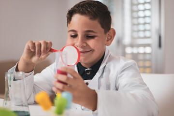 Boy having fun with chemistry lab in his living room. Scientific experiments at home for school homework. The little one looks at the liquid in the test tube with a magnifying glass.