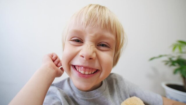 Cute 5 years old caucasian boy sits close to the camera and makes funny faces in it. A boy with fair hair and blue eyes smiles into the camera showing teeth