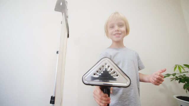 Smiling 5 years old caucasian boy looks into the camera and blows off steam from the professional garment steamer for clothes showing thumb up. Little helper helped to iron his clothes at home