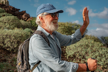 Active smiling senior man outdoors enjoying healthy lifestyle goes trekking in mountain environment. Senior bearded male in hat and backpack walking with stick greeting someone