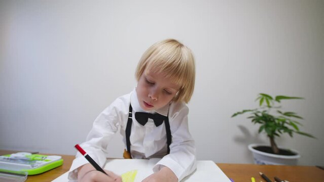 5 years old caucasian boy dressed in stylish tie bow and bright trousers draws in the big notebook with colorful pencils. Preschooler is busy drawing a picture in the album