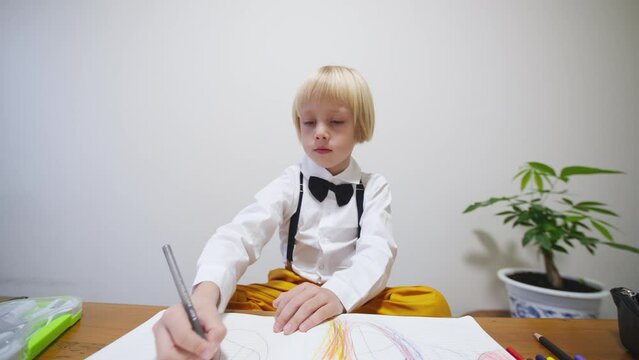 Zoom out. Preschooler is busy drawing a picture in the album. 5 years old caucasian boy dressed in stylish tie bow and bright trousers draws in the big notebook with colorful pencils. 
