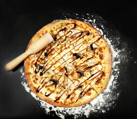 Appetizing fragrant Italian pizza with melted cheese and champignons on a black table with wooden kitchen utensils and scattered flour around; concept of world cuisines