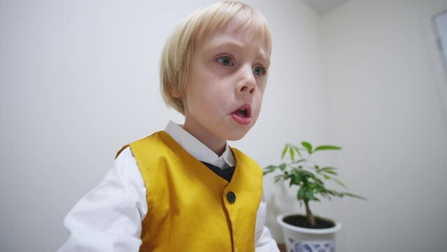 Portrait of a 5 years old caucasian boy in stylish bright waistcoat tie bow playing computer game at the laptop and making funny facs. The screen is reflected in his eyes