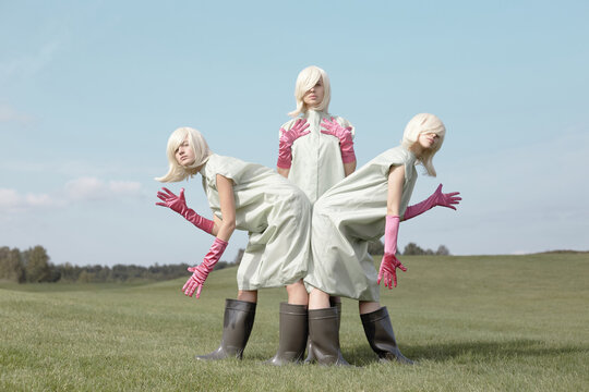 Conceptual shot of identical young blond women in clothing covers, pink gloves and rubber boots posing in bizarre postures on green field  