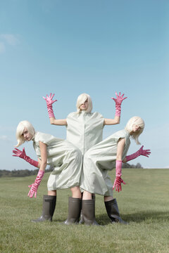 Conceptual photo of identical young women in weird costumes, pink gloves and rubber boots posing like robots outdoors on clear blue sky background  
