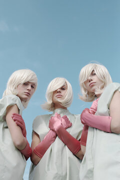 Conceptual photo of young alike blond women with pale skin wearing clothing cases and pink gloves looking at camera on clear blue sky background, low angle view 