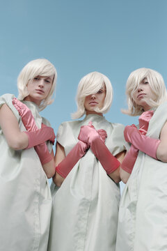 Conceptual shot of identical blond-haired girls with pale skin wearing garment covers and pink gloves looking at camera seriously against blue sky, low angle view  