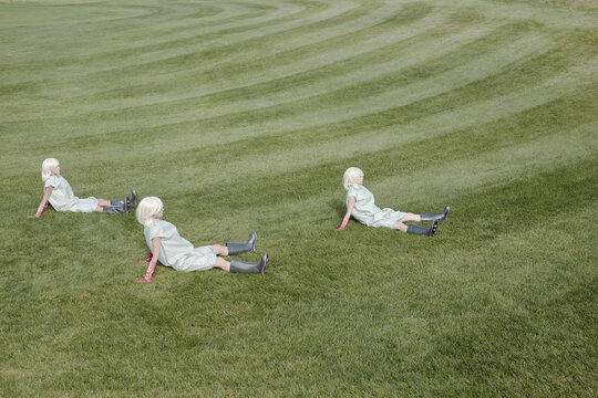 Conceptual shot of unrecognizable alike blond women wearing garment covers and  rubber boots sitting on green field and looking into the distance  