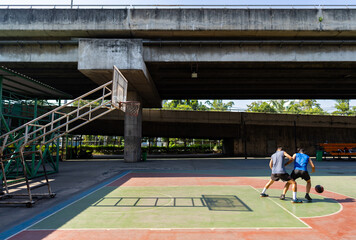 Obraz premium Two man athlete playing streetball match shooting and defense basketball on outdoors court together in sunny day. Sportsman do sport training basketball at street court under highway in the city.