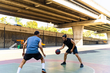 Fototapeta premium Two man athlete playing streetball match shooting and defense basketball on outdoors court together in sunny day. Sportsman do sport training basketball at street court under highway in the city.