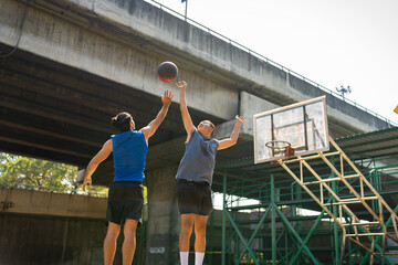 Two man athlete playing streetball match shooting and defense basketball on outdoors court together in sunny day. Sportsman do sport training basketball at street court under highway in the city.