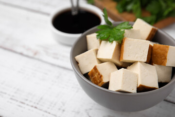 Bowl of smoked tofu cubes, soy sauce and parsley on white wooden table, closeup. Space for text