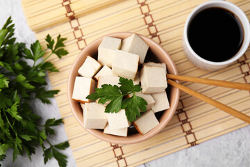 Bamboo mat with bowl of smoked tofu cubes, soy sauce and parsley on white textured table, flat lay