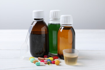 Bottles of syrup, measuring cup, dosing spoon and pills on white table. Cold medicine