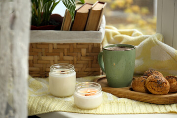 Burning aromatic candles, cup with hot drink and cookies on windowsill indoors. Autumn coziness