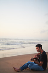 young latino male playing guitar and singing alone relaxed sitting on the beach shore at sunset