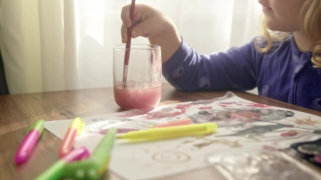 child rinses paint brush from red color in pink color water. Wash brush in transparent glass, paint with watercolors on paper at home. unrecognizable hand of girl cleans brush from paint