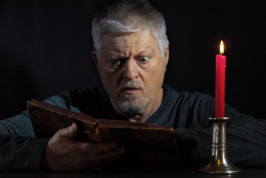 Portrait of an elderly man reading a book by candlelight. His gaze is intense. The background is black.