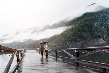 
A young sympathetic couple stands with an umbrella in their hands against the backdrop of a beautiful lake and mountains.