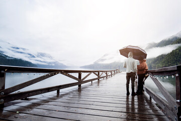 
A young sympathetic couple stands with an umbrella in their hands against the backdrop of a beautiful lake and mountains.