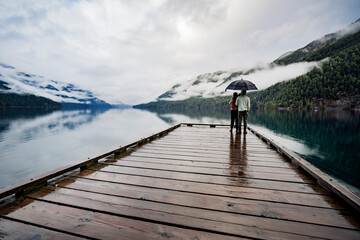 
A young sympathetic couple stands with an umbrella in their hands against the backdrop of a beautiful lake and mountains.
