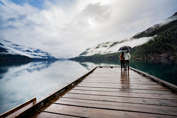 
A young sympathetic couple stands with an umbrella in their hands against the backdrop of a beautiful lake and mountains.