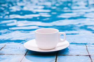 White cup of coffee by the edge of the pool, turquoise water background