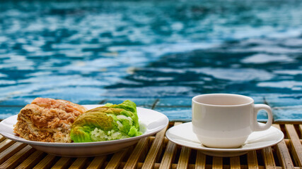 White cup of coffee and a plate of custard bread on the edge of the pool, turquoise water background