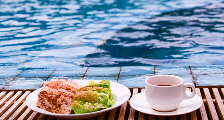 White cup of coffee with a plate of custard bread by the pool edge, blue water background