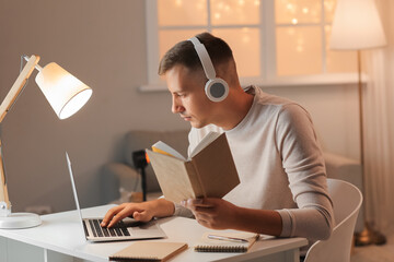 Young man with headphones, book and laptop studying online at home late in evening