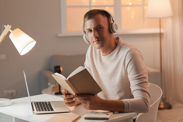 Young man with headphones, book and laptop studying online at home late in evening