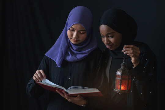 Beautiful multiethnic Muslim girl wearing hijab reading Quran, one holding Arab lamp, isolated on black background