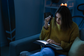 Young woman reading book with flashlight at home during blackout - obrazy, fototapety, plakaty