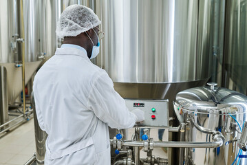 Rear view of specialist in protective clothing controlling the equipment of fermenting during his work at brewery