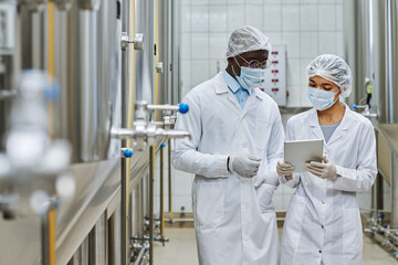 Two professional engineers in white coats and masks discussing something on digital tablet in workshop of brewery